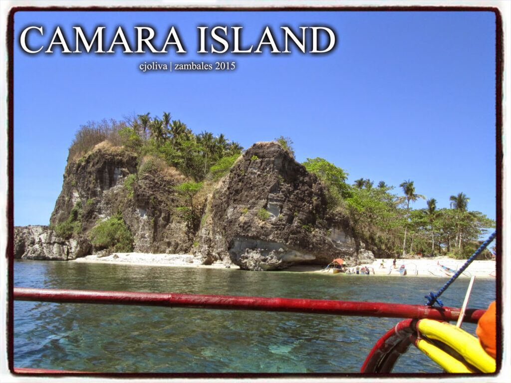 A view from a boat of Camara Island in Zambales, showing steep rocky cliffs, a small white sand beach, and palm trees under a clear blue sky.