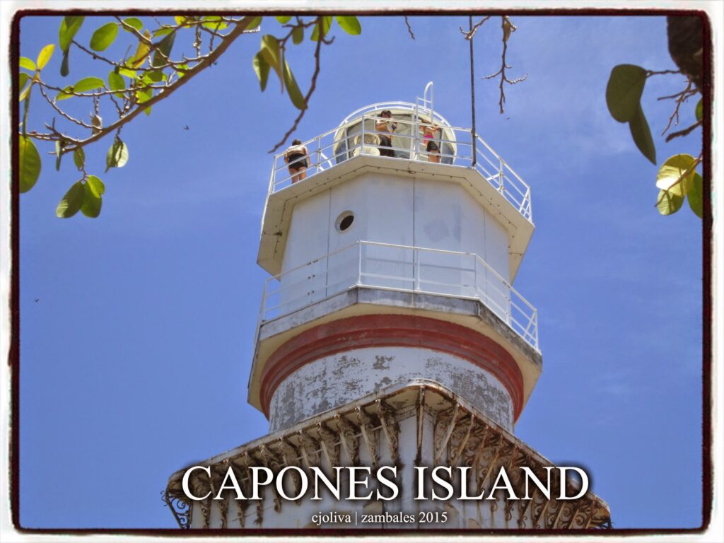 A low-angle view looking up at the white and red Faro de Punta Capones lighthouse on Capones Island, with people visible on the observation deck against a blue sky.