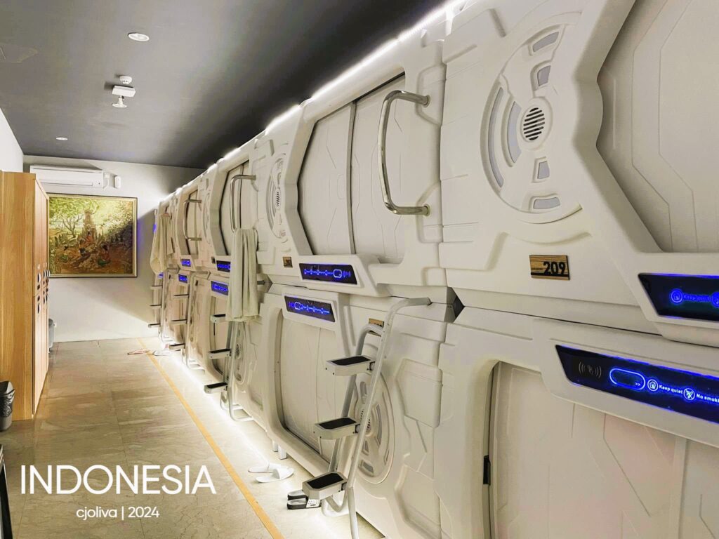 Modern capsule hotel interior in Indonesia with stacked pods, white panels, blue LED lights, ladders, and control panels.