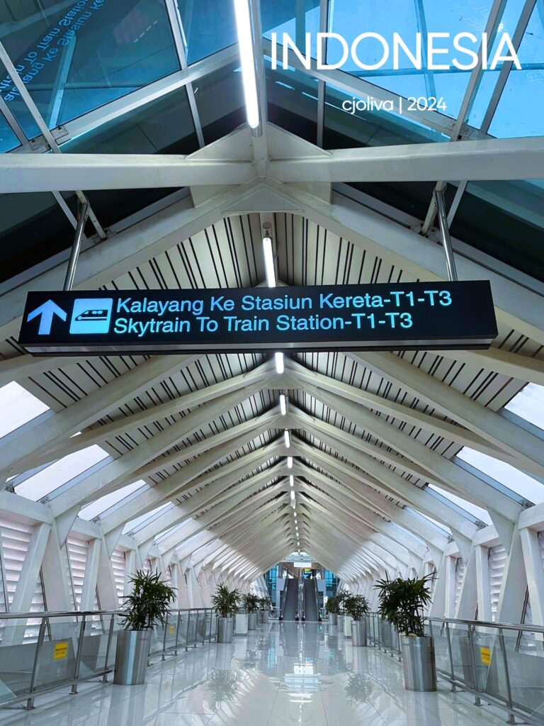 Modern enclosed pedestrian walkway at Soekarno–Hatta International Airport (CGK) in Jakarta, Indonesia, with glass panels, white beams, potted plants, and signage directing travelers to the skytrain between terminals T1–T3.