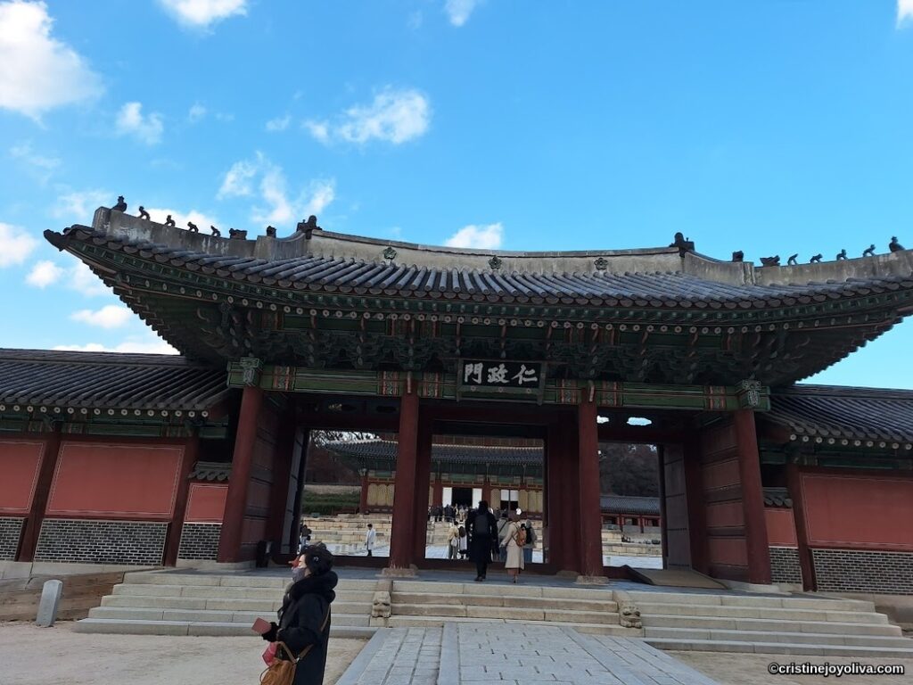 Tourists walking through Injeongmun Gate, the main entrance of Changdeokgung Palace in Seoul, showcasing traditional Joseon Dynasty architecture with an ornate roof and carvings.