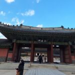 Tourists walking through Injeongmun Gate, the main entrance of Changdeokgung Palace in Seoul, showcasing traditional Joseon Dynasty architecture with an ornate roof and carvings.