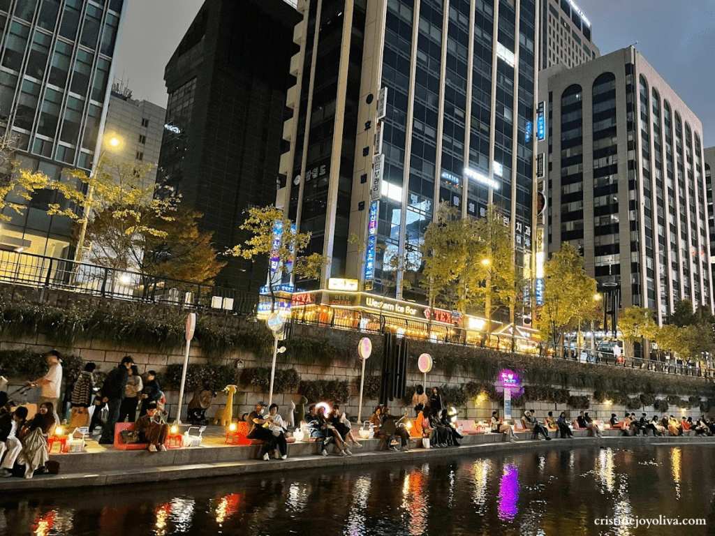 People sitting along the illuminated stone banks of Cheonggyecheon Stream at night, with city lights and skyscrapers reflecting in the water.