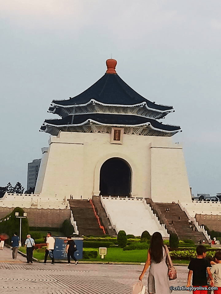 Chiang Kai‑shek Memorial Hall in Taipei, Taiwan, with white marble walls, a blue octagonal roof, red ornaments, a grand staircase, a landscaped plaza, and visitors walking.