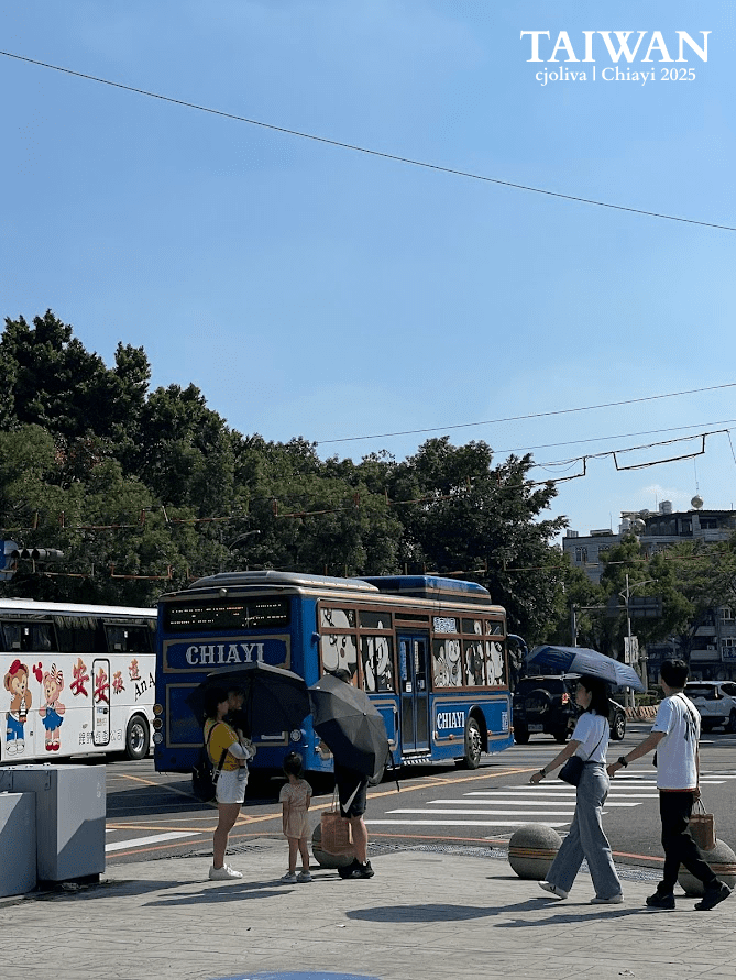Street scene in Chiayi, Taiwan, featuring a blue public bus decorated with cartoon characters and labeled “Chiayi,” with another illustrated bus in the background, pedestrians crossing the street, umbrellas for shade, and urban buildings under a sunny sky.