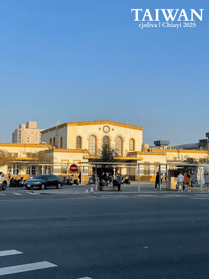 Front view of Chiayi Railway Station with arched windows, central clock, busy street with scooters, cars, and pedestrians under clear blue sky