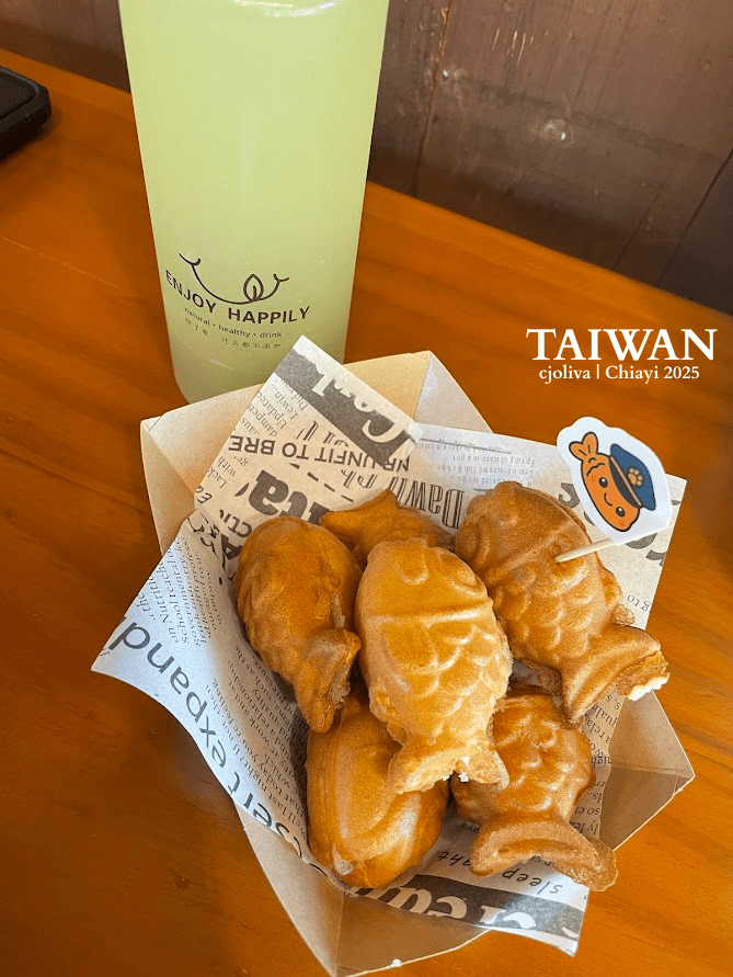 Fish-shaped taiyaki pastries in a paper-lined tray with creamy filling, accompanied by a light green bottled drink labeled “Enjoy Happily natural healthy drink,” on a wooden table in Chiayi, Taiwan.