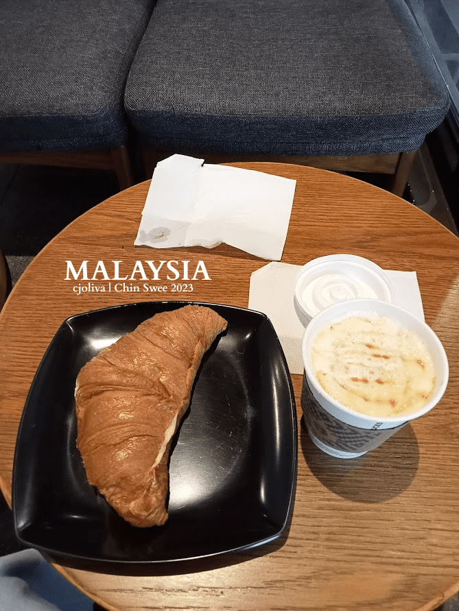 Croissant and coffee served at a café in Chin Swee, Malaysia, on a wooden table with cushioned seating in the background.