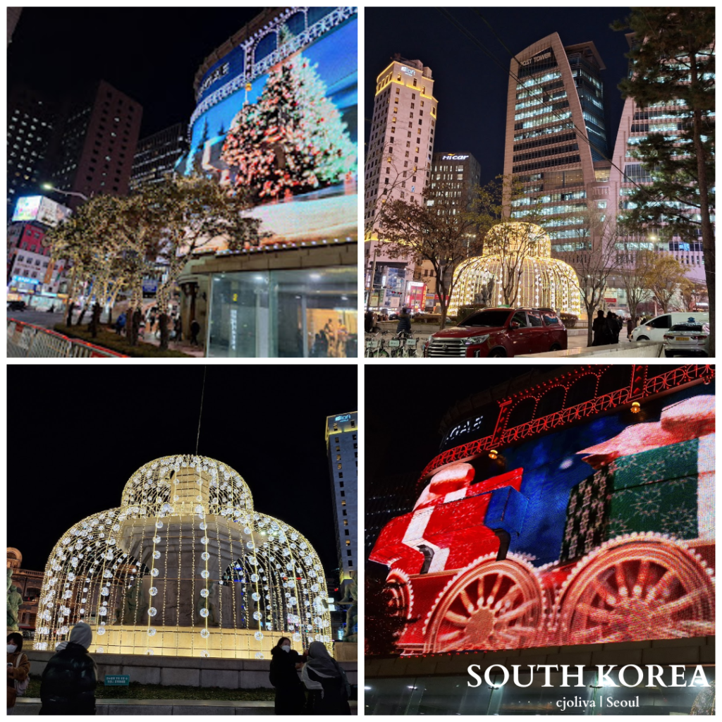 A brightly lit street in Seoul featuring large digital displays with Christmas themes, including a Christmas tree and a train, during nighttime.