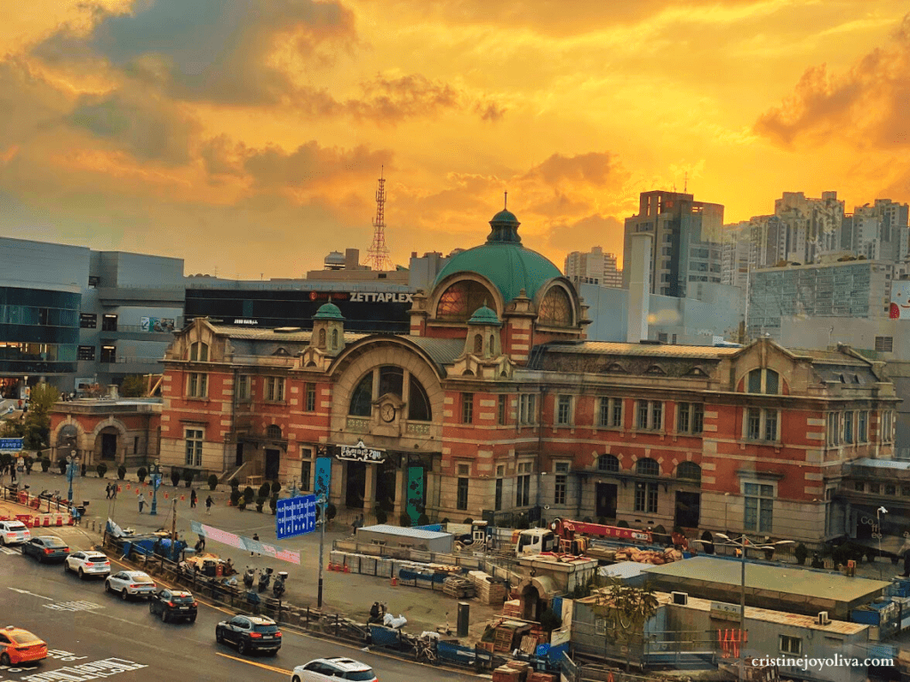 The historic red-brick facade and green dome of the old Seoul Station building, now Culture Station Seoul 284, set against a vibrant orange and yellow sunset sky with city traffic in the foreground.