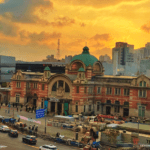 The historic red-brick facade and green dome of the old Seoul Station building, now Culture Station Seoul 284, set against a vibrant orange and yellow sunset sky with city traffic in the foreground.