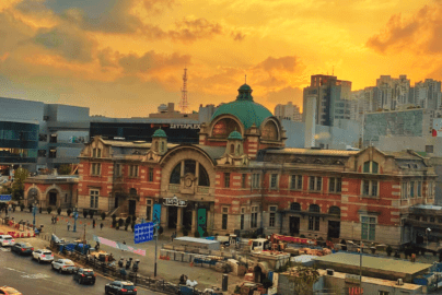 The historic red-brick facade and green dome of the old Seoul Station building, now Culture Station Seoul 284, set against a vibrant orange and yellow sunset sky with city traffic in the foreground.