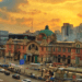 The historic red-brick facade and green dome of the old Seoul Station building, now Culture Station Seoul 284, set against a vibrant orange and yellow sunset sky with city traffic in the foreground.