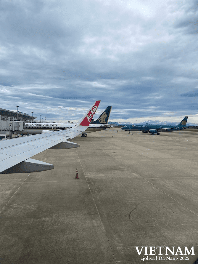 A view from an airplane window at Da Nang International Airport, showing a Vietnam Airlines plane, a Singapore Airlines tail, and an AirAsia wing on the tarmac under a cloudy sky.