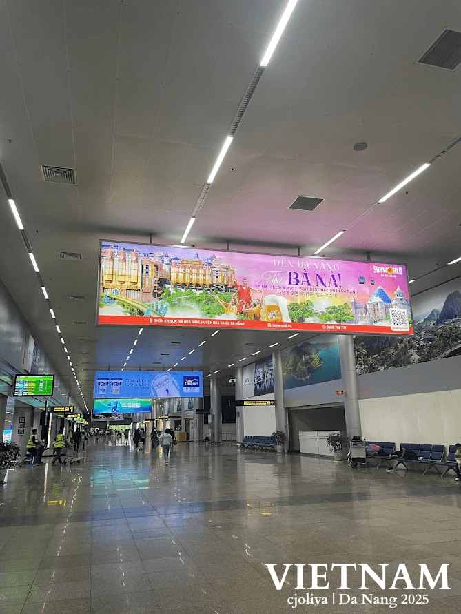 Interior of Da Nang airport terminal in Vietnam showing polished floors, overhead lighting, directional signage, seating areas, travelers, and a large billboard promoting Ba Na Hills tourist destination.