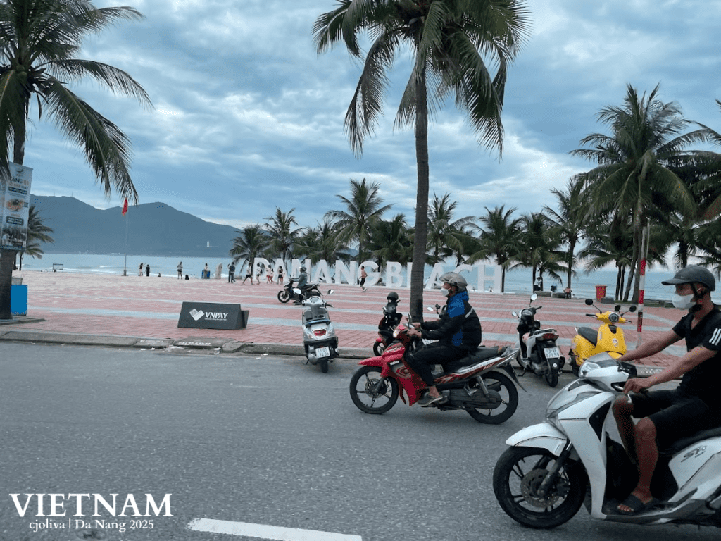 A coastal view in Da Nang, Vietnam, featuring palm trees, a white 'DA NANG BEACH' sign, and locals on motorbikes driving past the sandy shore under a cloudy sky.