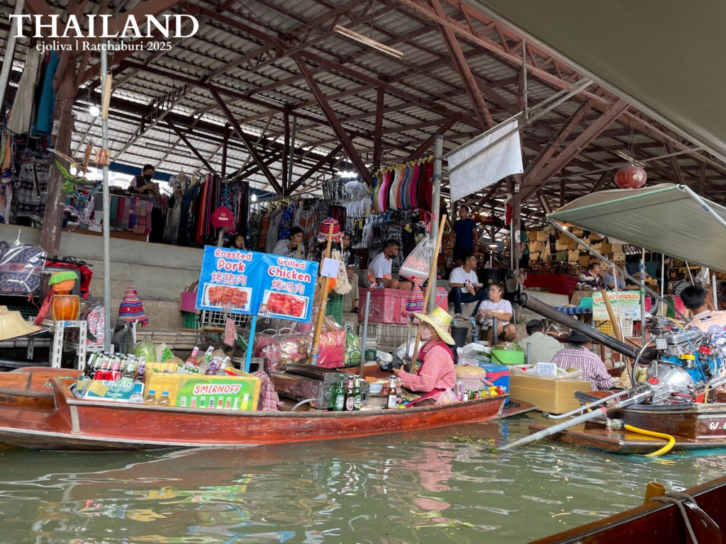 A wooden boat at Damnoen Saduak Floating Market in Ratchaburi, Thailand, serving grilled chicken, roasted pork, and cold beverages under a large covered market structure.
