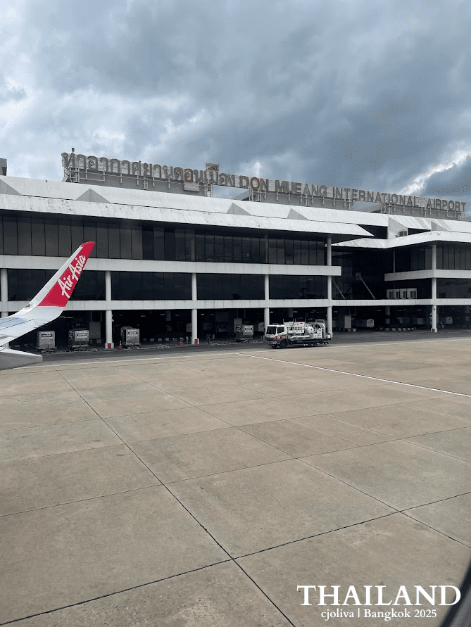 An exterior view of the Don Mueang International Airport terminal building in Bangkok, Thailand, showing the airport's name in Thai and English on the roof.