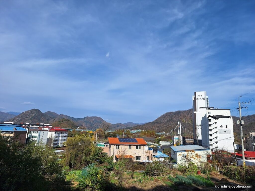 Rural town near Gapyeong Station in South Korea, with colorful rooftops, solar panels, and a suspension bridge against forested mountains.
