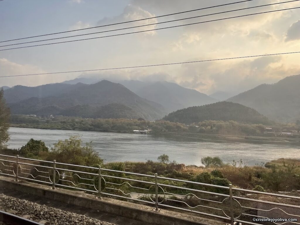 Scenic river and mountain view from railway tracks in Gapyeong, South Korea, with sunlight filtering through clouds.