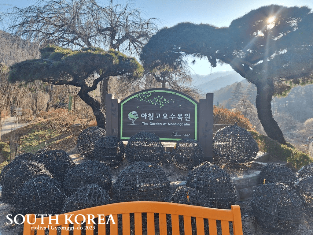 Entrance sign at the Garden of the Morning Calm in Gyeonggi-do, South Korea, surrounded by pruned trees, wire sculptures, and a mountain backdrop.