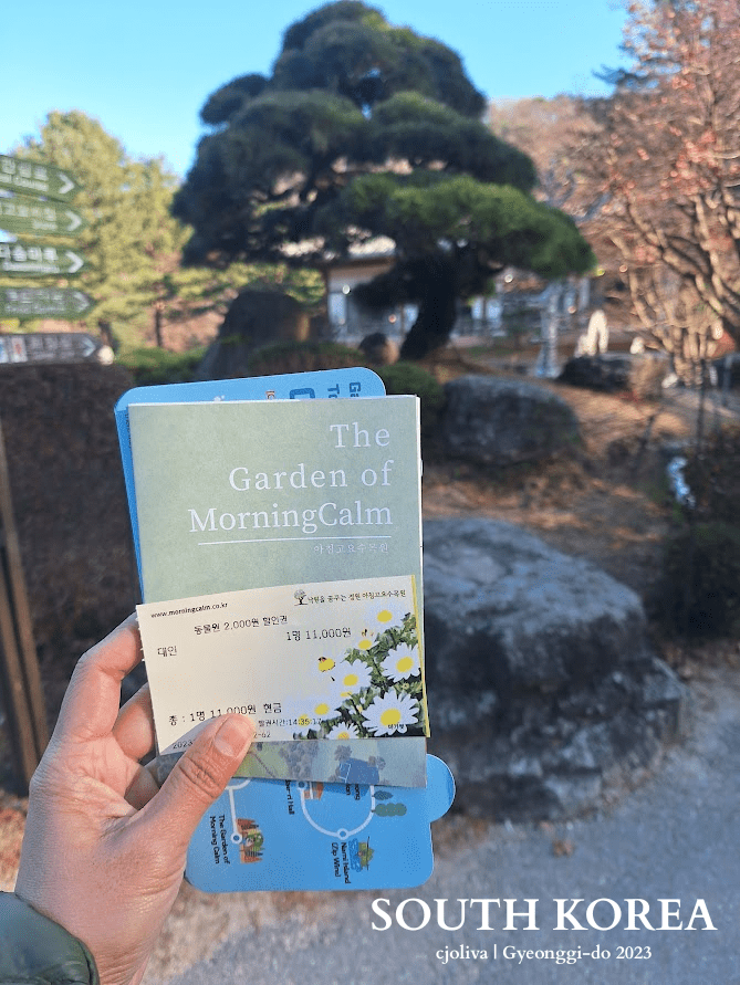 Visitor holding ticket and brochure for the Garden of the Morning Calm in Gyeonggi-do, South Korea, with a scenic garden background.