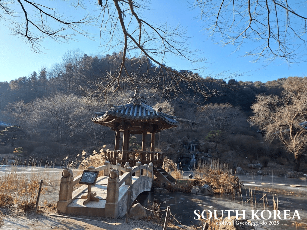 Traditional Korean pavilion on arched stone bridge over frozen pond in Guri-si, Gyeonggi-do, South Korea, surrounded by reeds and winter trees.