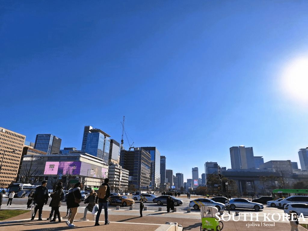 A wide-angle view of Gwanghwamun Square in Seoul featuring modern glass skyscrapers, a busy street with white and orange taxis, and pedestrians under a bright, clear blue sky.