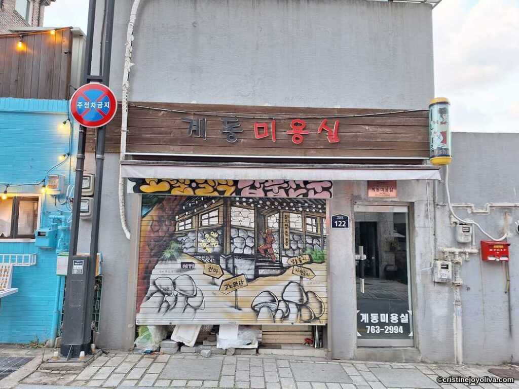 Exterior of Gyedong Beauty Salon in Seoul with painted mural on shutter, red signage, and traditional-style cartoon interior scene.