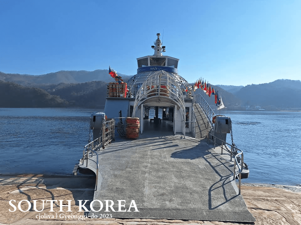 Docked ferry with boarding ramp in Gyeonggi-do, South Korea, surrounded by forested hills and calm water under a clear sky.