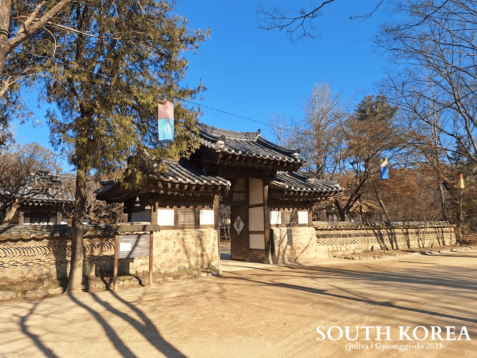 Traditional Korean gate with tiled roof, stone walls, and colorful lanterns hanging from trees at Korean Folk Village in Gyeonggi-do, South Korea, 2023