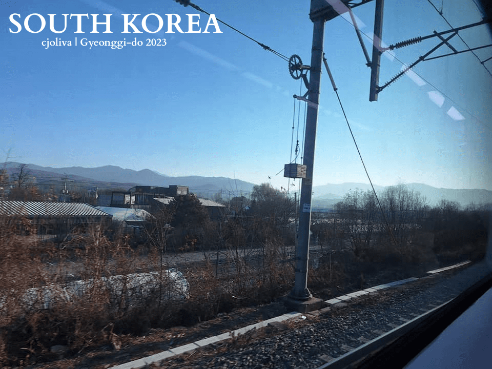 Scenic train journey through Gyeonggi-do, South Korea with railway tracks, rooftops, and distant mountains under a clear sky.