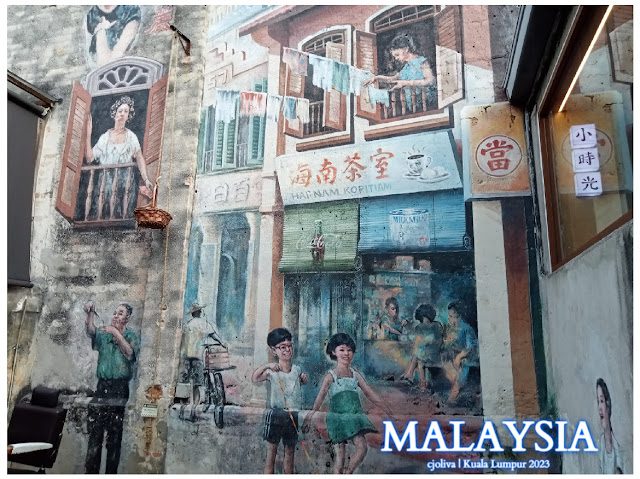 Street mural in Kuala Lumpur, Malaysia, depicting nostalgic daily life with a Hainan Kopitiam shopfront, children playing, clotheslines, a bicycle, and traditional cultural scenes.