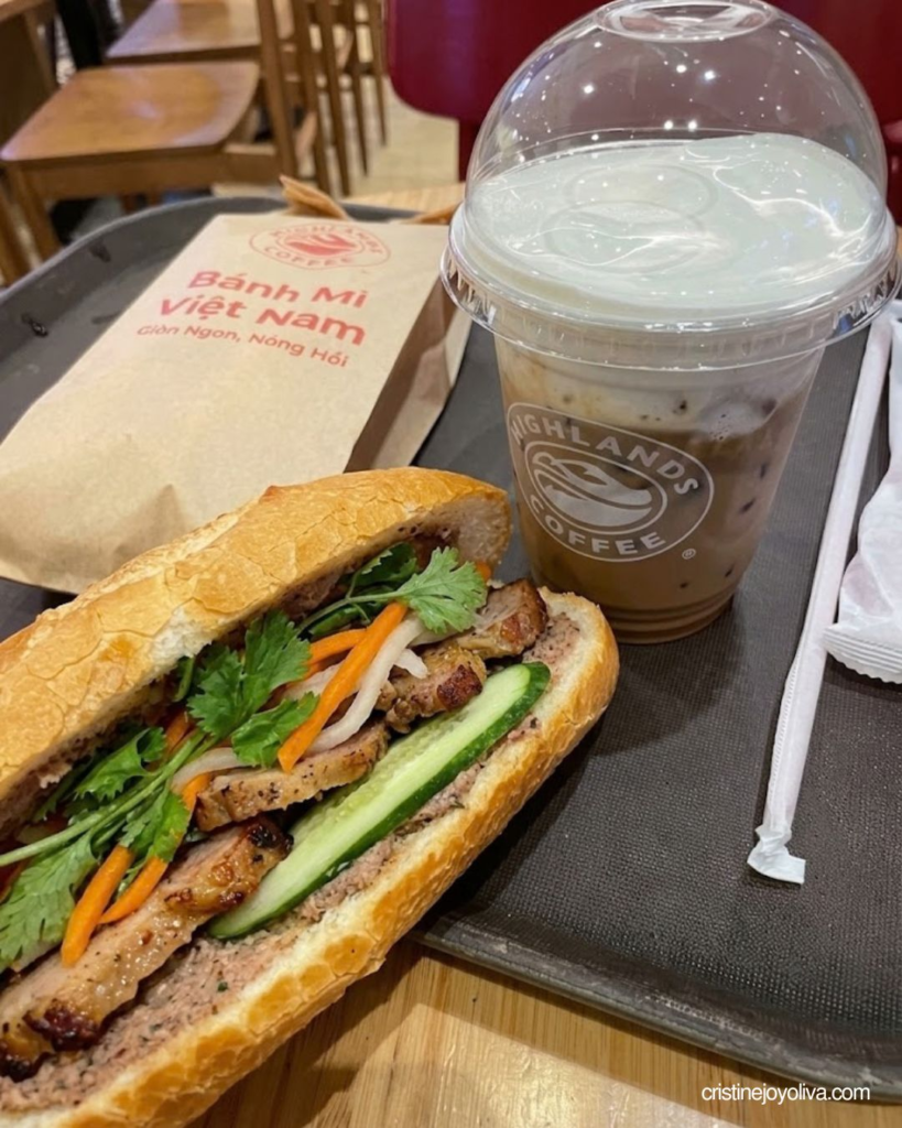 A close-up of an open-cut savory bánh mì sandwich with grilled meat, cucumbers, and cilantro beside an iced coffee from Highlands Coffee.