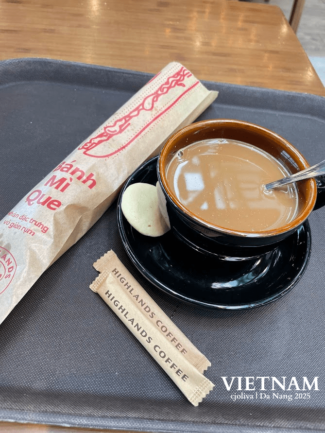 A black cup of Vietnamese milk coffee served with a small cookie and a paper-wrapped Bánh Mì Que breadstick on a tray at Highlands Coffee.