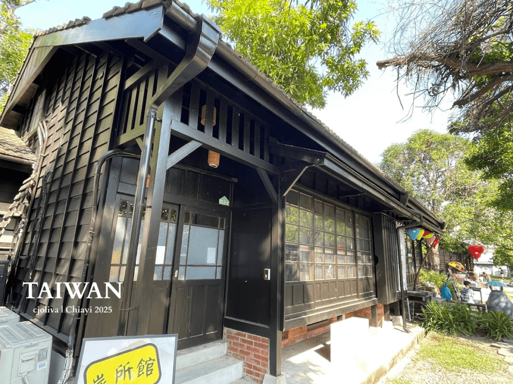 Traditional wooden building at Hinoki Village in Chiayi, Taiwan, with dark wood paneling, a tiled roof, colorful lanterns, landscaped greenery, and visitors exploring the cultural site.
