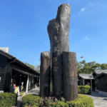 Large wooden sculpture with vertically arranged Chinese characters reading “Hinoki Village” in an outdoor plaza, surrounded by traditional black wooden buildings under a clear blue sky.