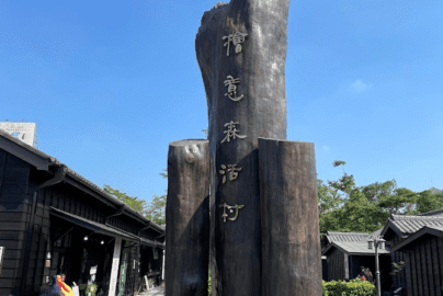 Large wooden sculpture with vertically arranged Chinese characters reading “Hinoki Village” in an outdoor plaza, surrounded by traditional black wooden buildings under a clear blue sky.