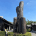 Large wooden sculpture with vertically arranged Chinese characters reading “Hinoki Village” in an outdoor plaza, surrounded by traditional black wooden buildings under a clear blue sky.
