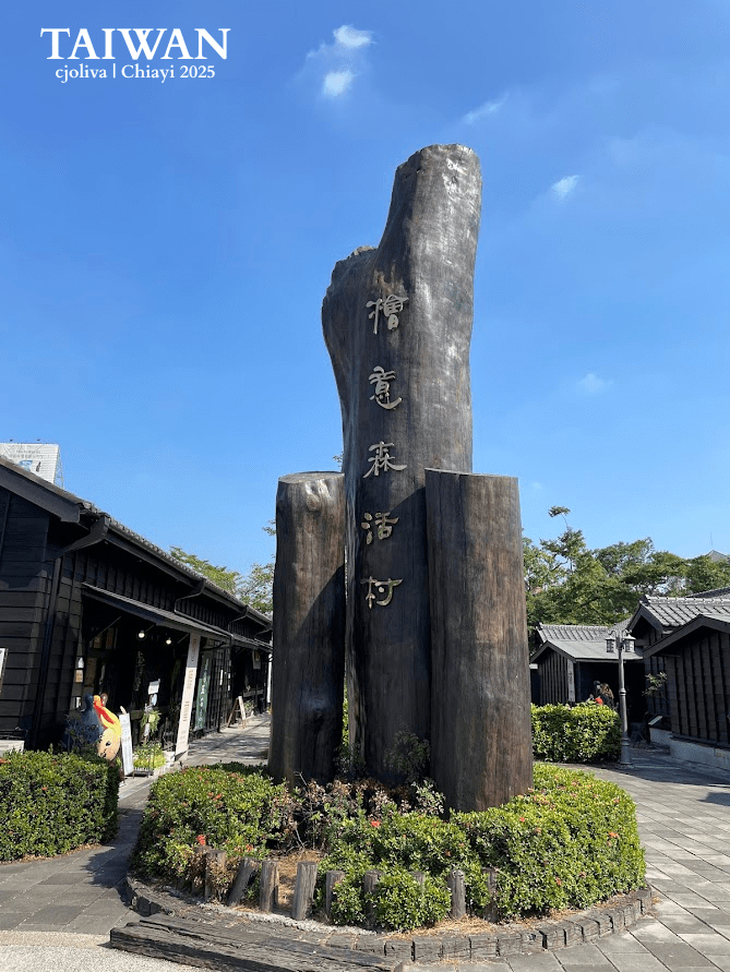 Large wooden sculpture with vertically arranged Chinese characters reading “Hinoki Village” in an outdoor plaza, surrounded by traditional black wooden buildings under a clear blue sky.