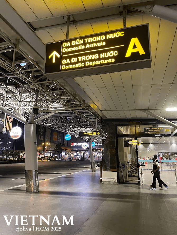 A Vietnam airport terminal scene in Ho Chi Minh City showing bilingual signage for Domestic Arrivals and Departures with a large yellow “A,” modern ceiling design, brand advertisements, and passengers walking.