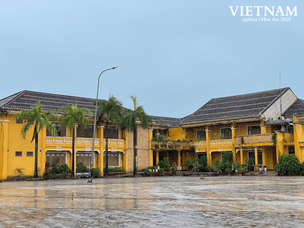 Vibrant yellow colonial buildings in Hoi An Ancient Town, Vietnam, after rain, with a wet courtyard, palms, and blue sky.