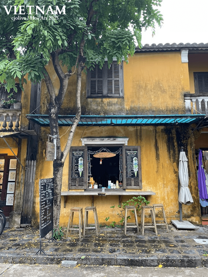 Rustic yellow ice cream shop in Hoi An, Vietnam, with wooden shutters, a blue awning, stools, and menu boards under a shady tree.