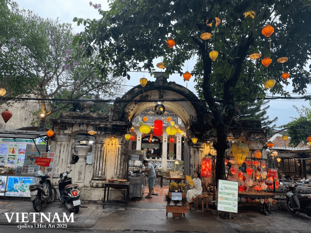 Weathered stone archway in Hoi An, Vietnam, with colorful lanterns, motorbikes, and signs for money exchange and tours, with a person in traditional attire seated nearby.