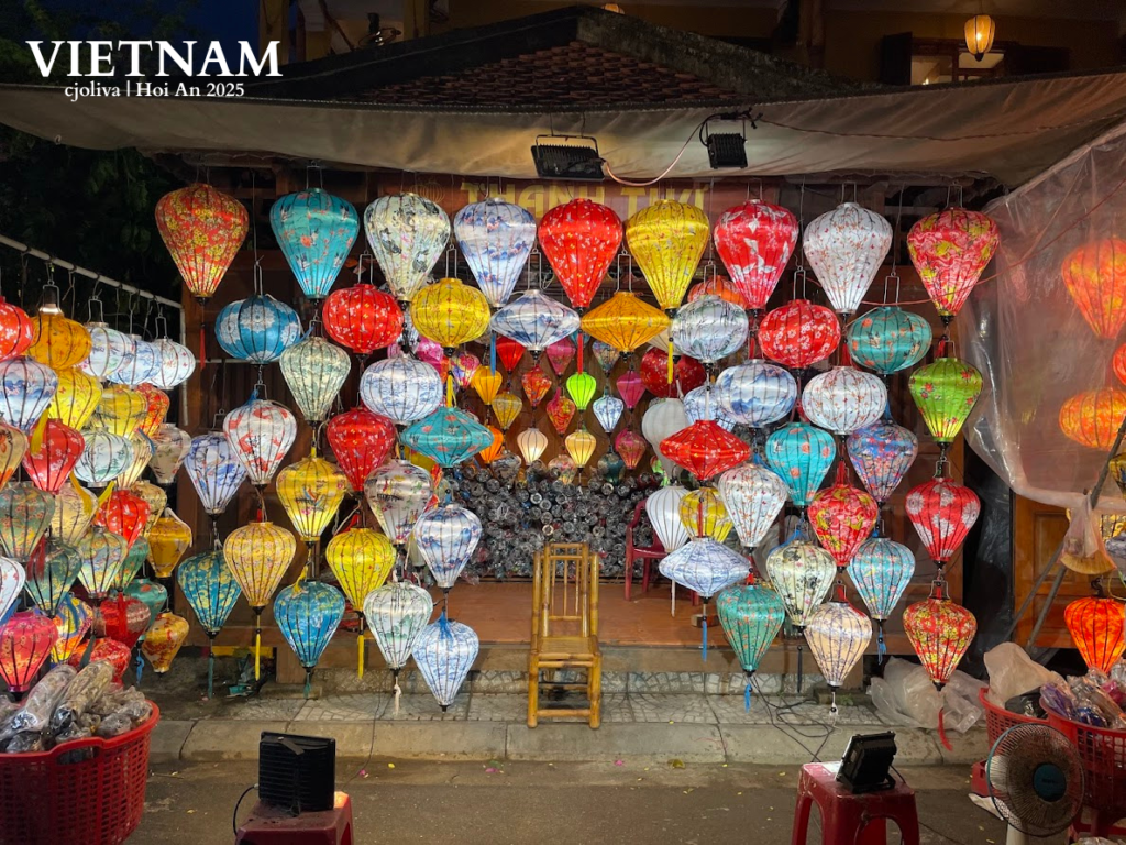 Colorful illuminated lanterns hanging in front of a shop in Hội An, Vietnam, with stools, baskets, and a wooden chair in the market setting.