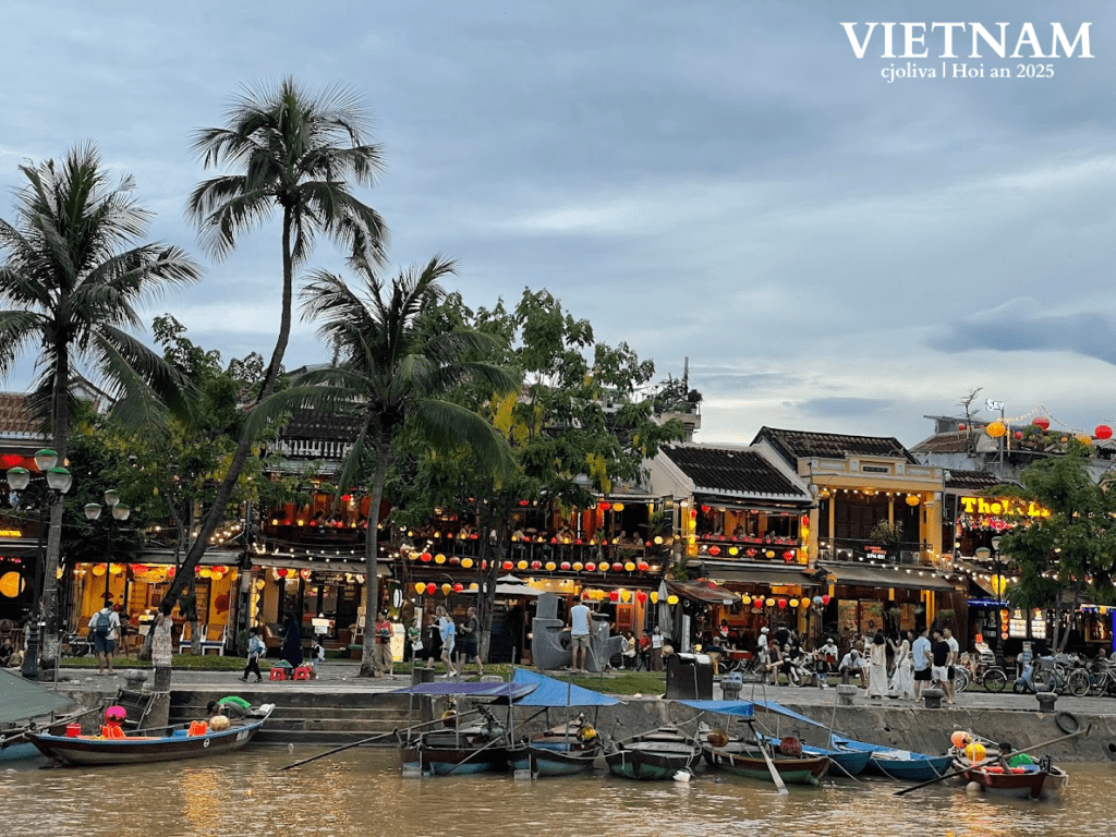 Riverside in Hoi An, Vietnam, with small boats, palm trees, traditional lantern-lit buildings, and people gathering along the promenade under an overcast sky.