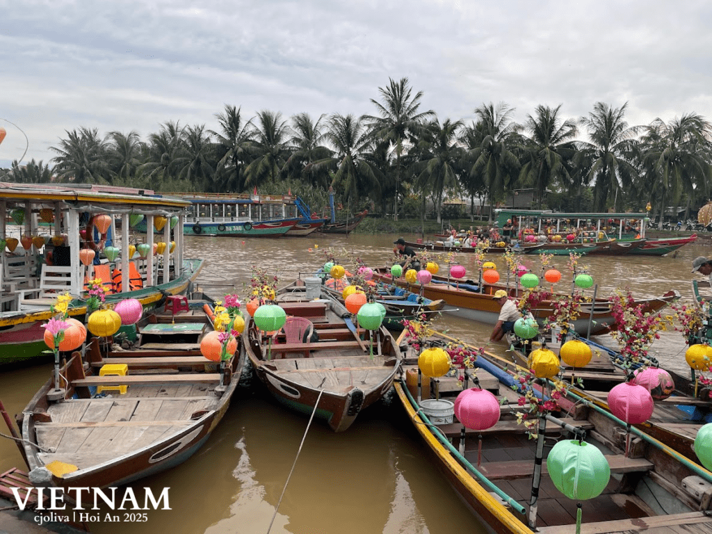 Traditional wooden boats decorated with colorful lanterns and flowers on the river in Hoi An, Vietnam, with palm trees along the riverbank under an overcast sky.