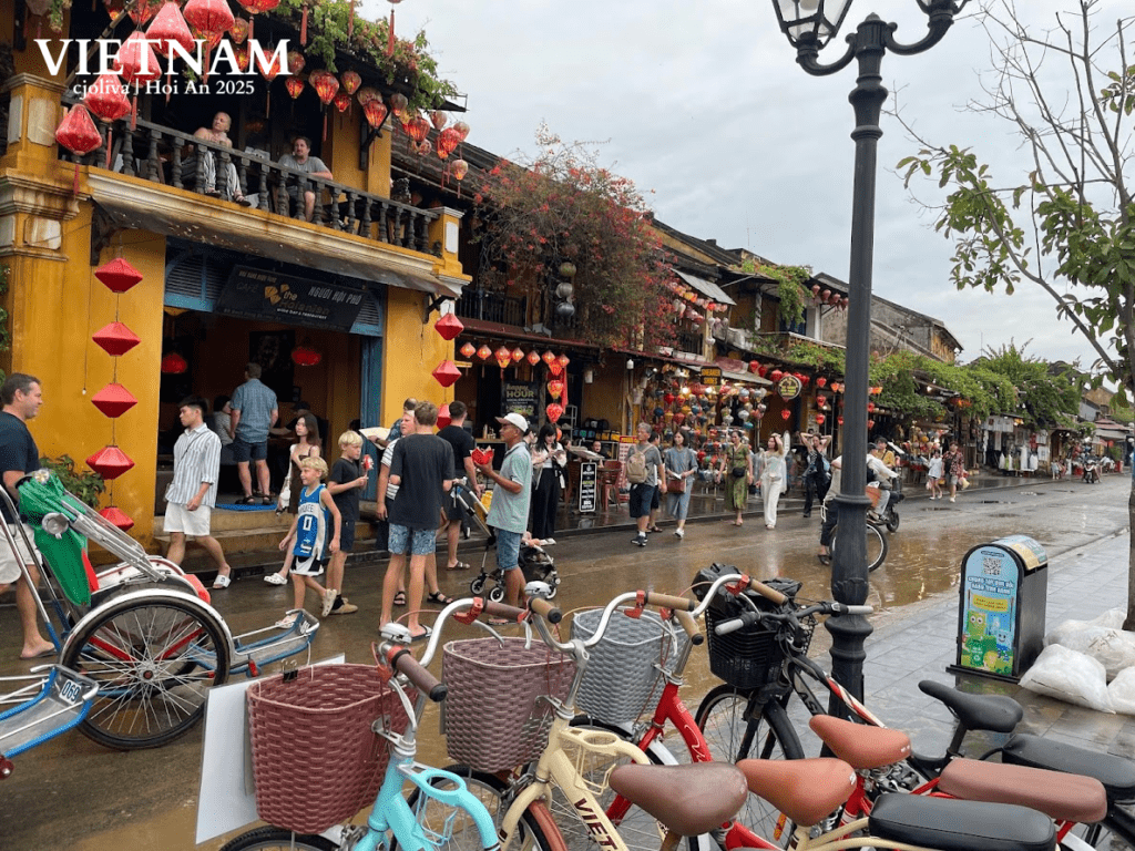 Bustling street in Hoi An, Vietnam, with yellow heritage buildings, red lanterns, flowering plants, bicycles, and crowds of people during a festival.