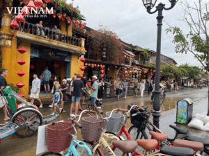 Bustling street in Hoi An, Vietnam, with yellow heritage buildings, red lanterns, flowering plants, bicycles, and crowds of people during a festival.