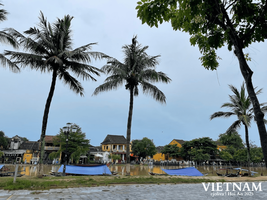 Scenic view of Hoi An, Vietnam, with yellow heritage buildings, palm trees, and boats along the riverbank under an overcast sky.